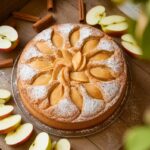 A freshly baked apple cake topped with powdered sugar and cinnamon on a rustic wooden table.